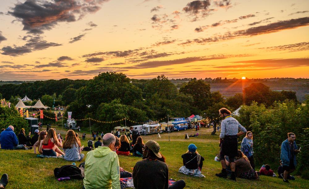 Festival audience sat on hill enjoying the sunset through the trees.