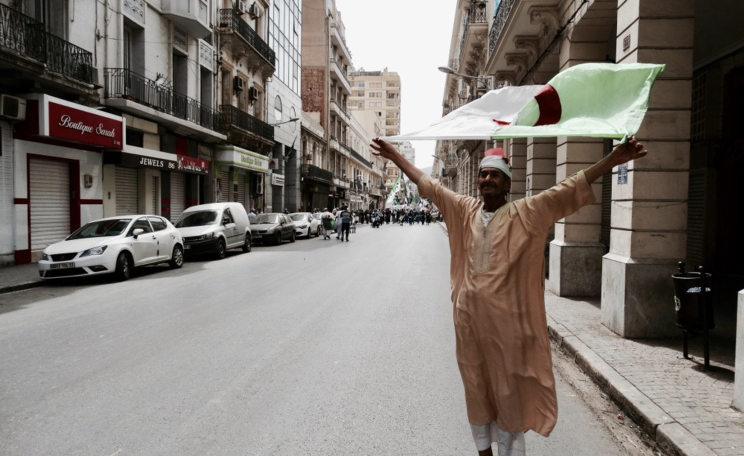 A man stands waving Algerian flag on the streets of Oran as a mass  of protestors approach. 